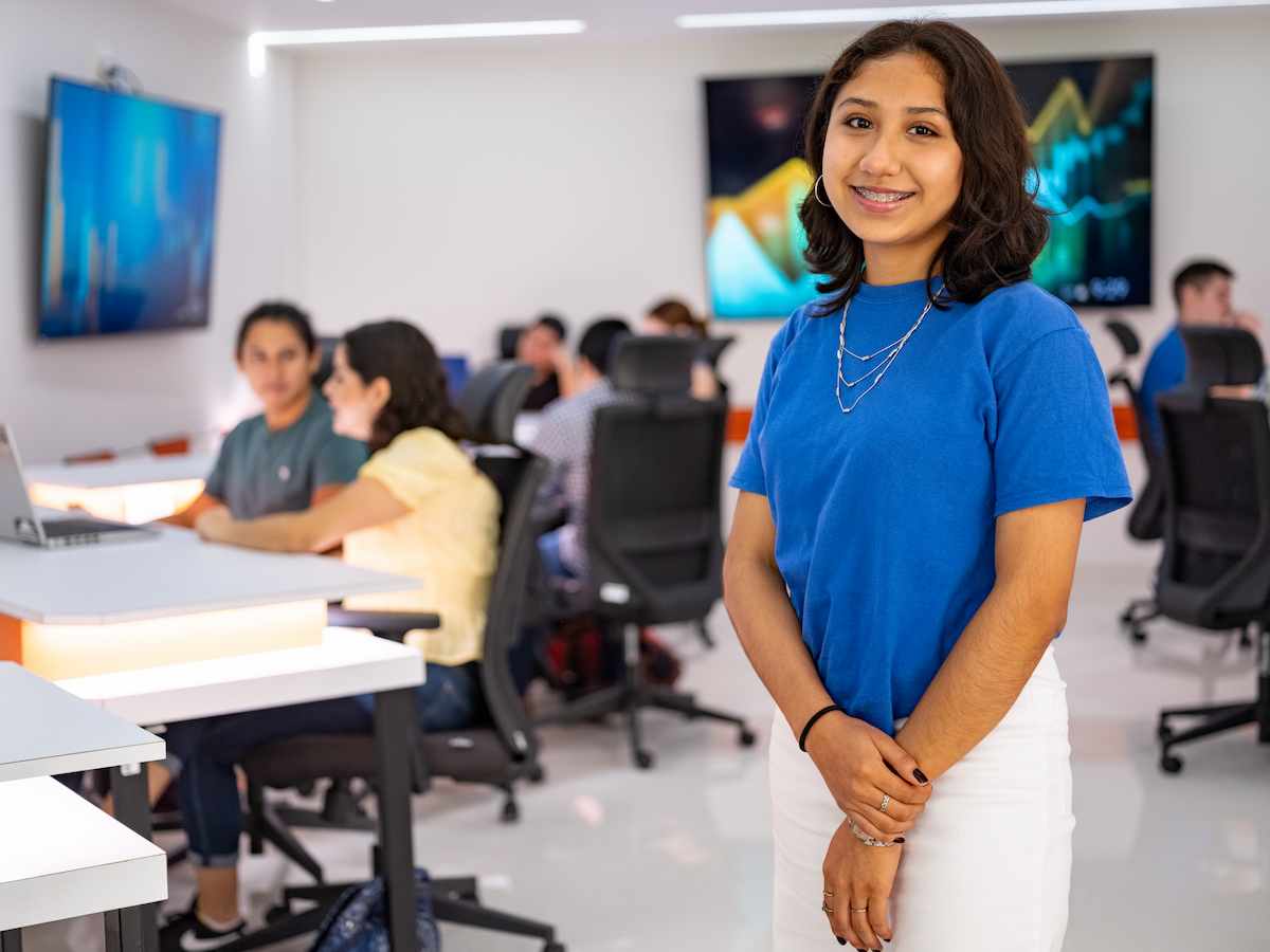 Estudiante con blusa azul y falda blanca sonríe al frente de un aula moderna con estudiantes trabajando en computadoras al fondo.